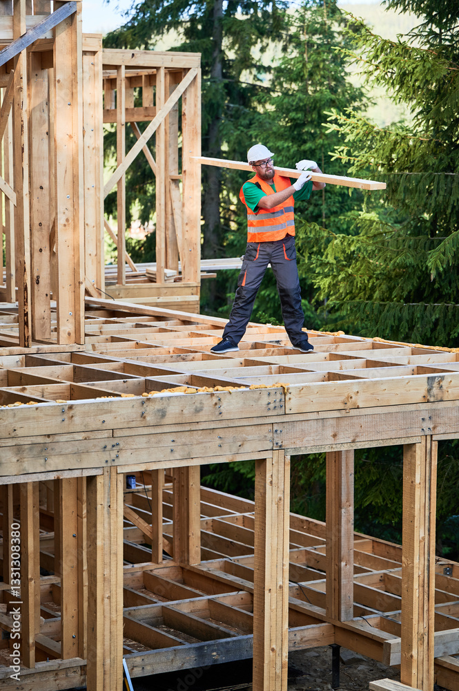 Carpenter building wooden frame house near the forest. Bearded man with spectacles holding large board on his shoulder, dressed in work clothes and helmet. Concept of modern, sustainable construction. Carpenter building wooden frame house near the forest. Bearded man with spectacles holding large board on his shoulder, dressed in work clothes and helmet. Concept of modern, sustainable construction.