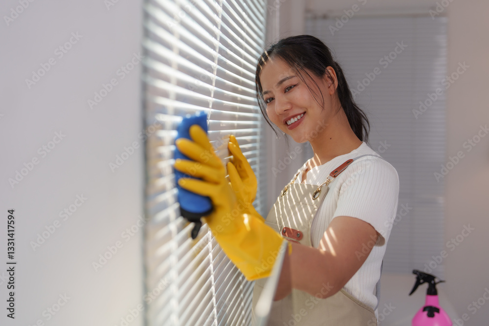 Young, smiling asian cleaning lady wearing yellow gloves and an apron, diligently washing window blinds with a blue sponge and detergent from a spray bottle, ensuring a spotless finish Young, smiling asian cleaning lady wearing yellow gloves and an apron, diligently washing window blinds with a blue sponge and detergent from a spray bottle, ensuring a spotless finish