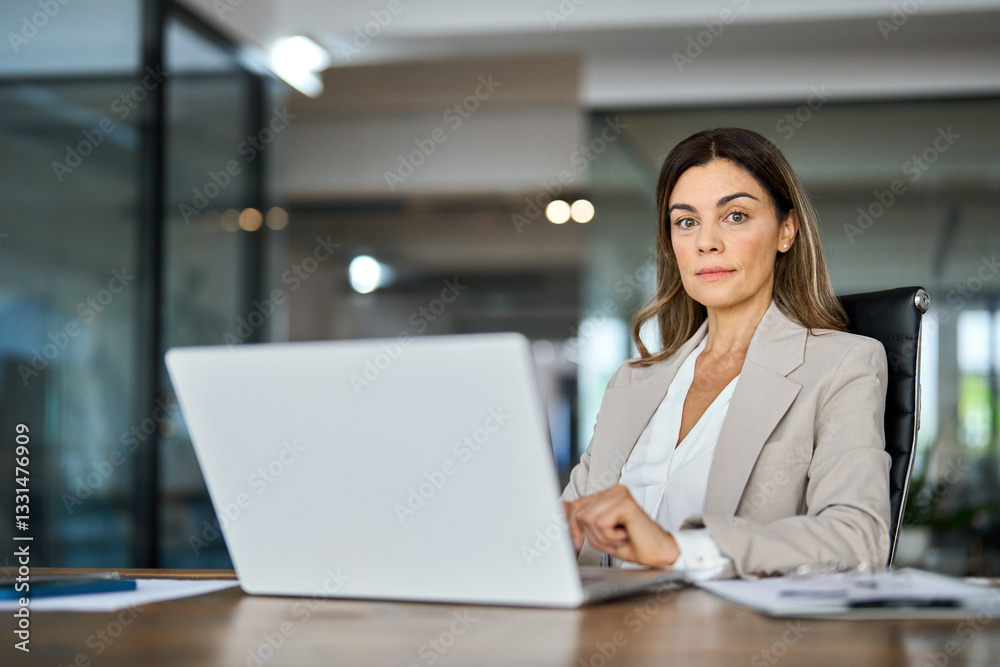 Confident mature middle aged professional business woman boss, female manager executive looking at camera at workplace, working on laptop computer technology in office sitting at desk, portrait. Confident mature middle aged professional business woman boss, female manager executive looking at camera at workplace, working on laptop computer technology in office sitting at desk, portrait.
