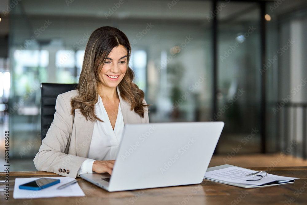 Smiling happy busy mature middle aged professional business woman manager executive wearing suit looking at laptop computer technology in office working on digital project sitting at desk.