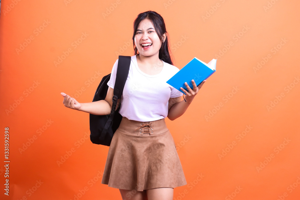 Happy student with backpack holding a book in her hand wearing a white t shirt and a skirt is isolated on orange background Happy student with backpack holding a book in her hand wearing a white t shirt and a skirt is isolated on orange background