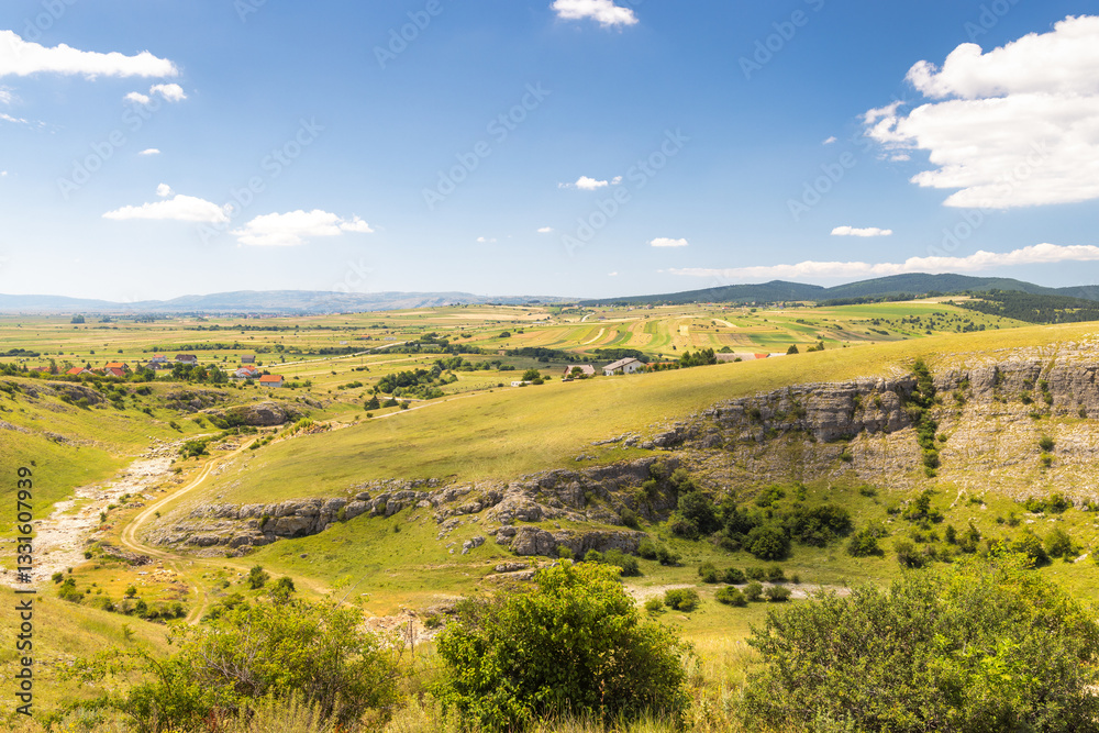 Fototapeta premium View of country in Bosnia and Herzegovina, Europe. Scenic landscape panorama of rolling hills and fields under a blue sky with scattered clouds, offering a peaceful, natural countryside vista.