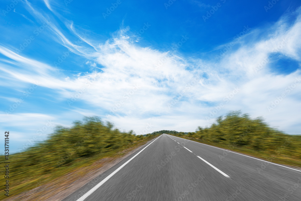 A dynamic image of an open highway surrounded by greenery, with a motion blur effect on the sides, leading into the horizon under a bright blue sky with wispy clouds.
