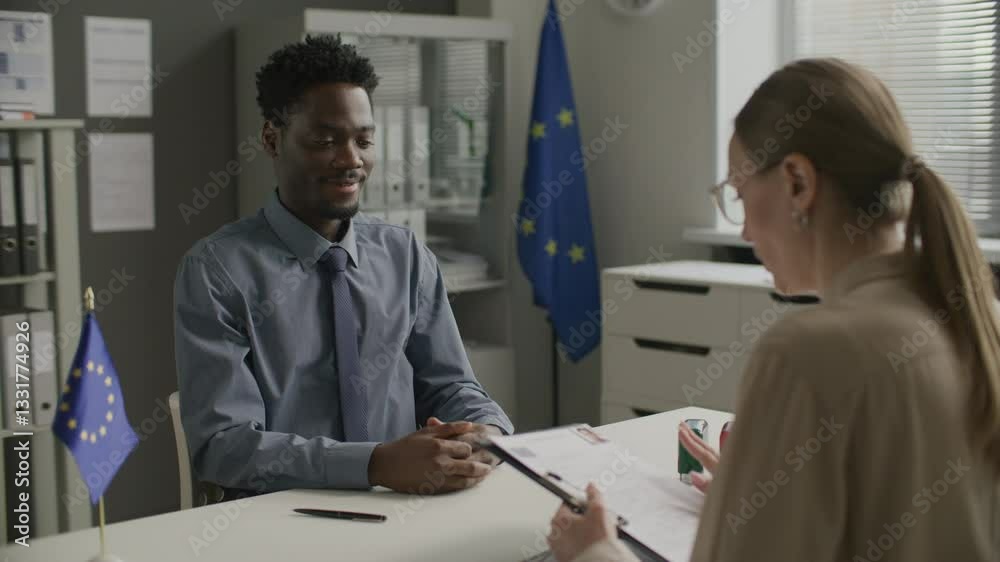 Young African American man passing application form to female visa officer, she checking documents and interviewing him at European Union embassy