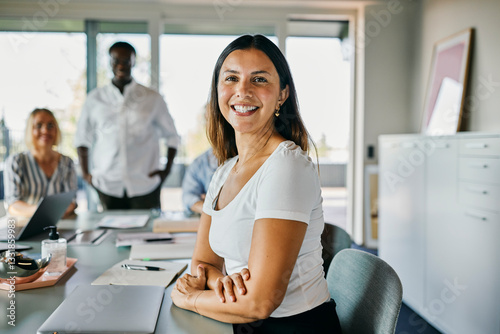 Confident smiling businesswoman leading a successful team meeting in modern office. Behind her stands a colleague, leaning over the table.