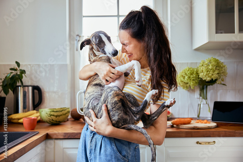 Joyful woman holding whippet puppy standing in kitchen
