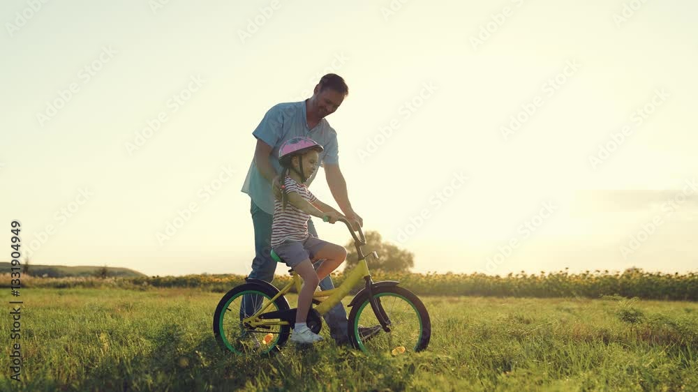 Smiling father teaches little girl to ride bicycle in summer field. Preschooler girl with helmet rides bicycle with loving father in park. Happy father and girl plays with bicycle in meadow