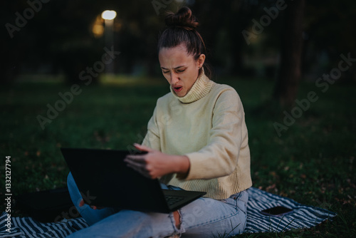 Obraz na plátně A young girl sitting on a blanket outdoors, expressing frustration while using her laptop during an evening setting