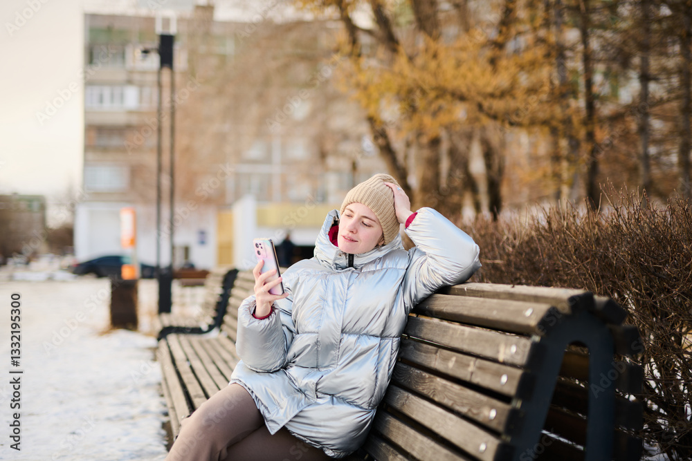 Fototapeta premium Portrait young Caucasian girl using smartphone, looking at mobile phone screen on winter walk in park