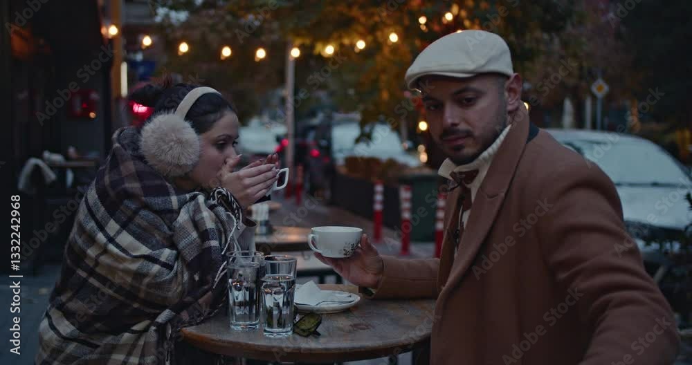 A loving couple enjoys a warm drink together at an outdoor cafe on a chilly autumn evening in the city. A loving couple enjoys a warm drink together at an outdoor cafe on a chilly autumn evening in the city.