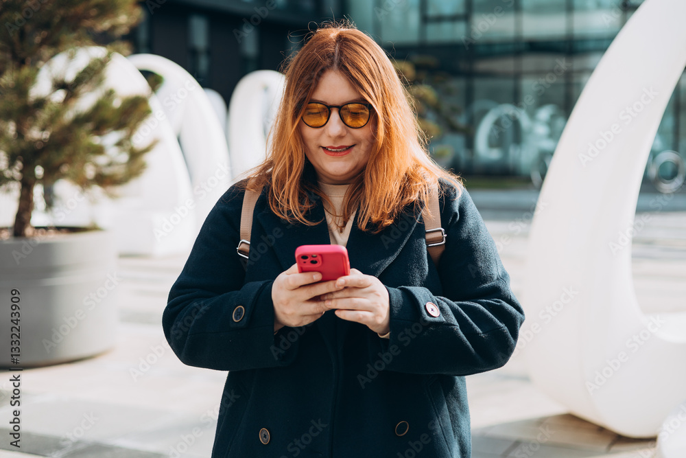 Happy woman chatting on phone and relaxing outdoors. Urban, people concept. Smiling 30s redhead woman text messaging on smart phone at sunny day on the city street, spring time