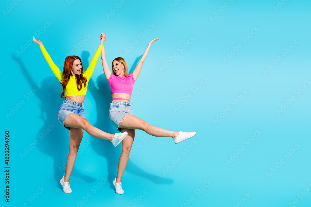 Two cheerful young girlfriends with red hair and blonde hair in bright summer outfits celebrating against turquoise background Two cheerful young girlfriends with red hair and blonde hair in bright summer outfits celebrating against turquoise background
