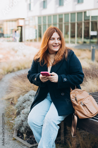 Happy woman sitting on the bench and chatting on phone outdoors. Urban, people concept. Smiling 30s redhead woman text messaging on smart phone at sunny day on the city street, spring time