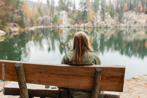 Lonely 30s blonde woman is sitting on the bench of a forest lake. Autumn landscape, calmness, tranquility, travel concept. A woman on the bank of a lake is enjoying the nature. High quality photo.