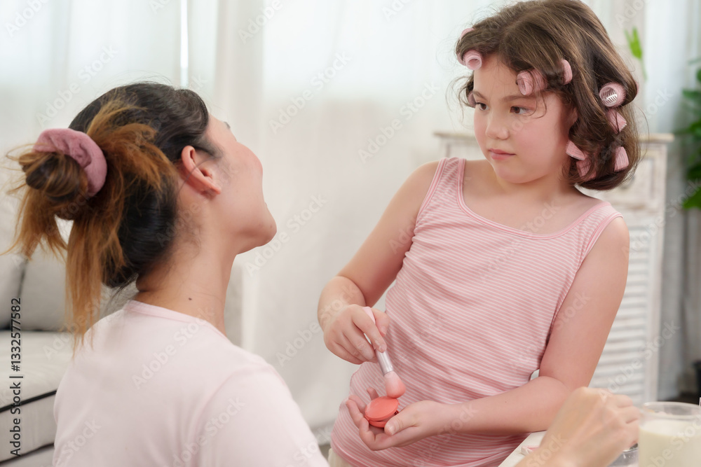 Caucasian girl holding blush powder while looking at Asian mother. Beauty lesson and playful makeup learning moment between mother and daughter in a warm and loving home environment. Caucasian girl holding blush powder while looking at Asian mother. Beauty lesson and playful makeup learning moment between mother and daughter in a warm and loving home environment.