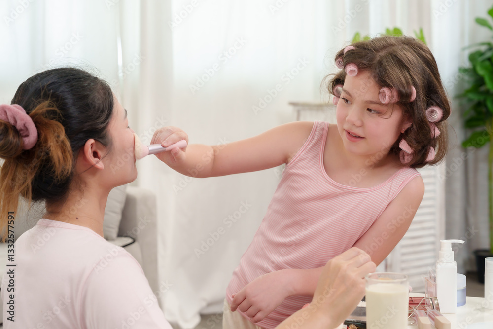 Fototapeta premium Caucasian girl playfully applying makeup on Asian mother’s face with a brush. Warm and happy family bonding moment, learning beauty techniques together in a cozy home setting.