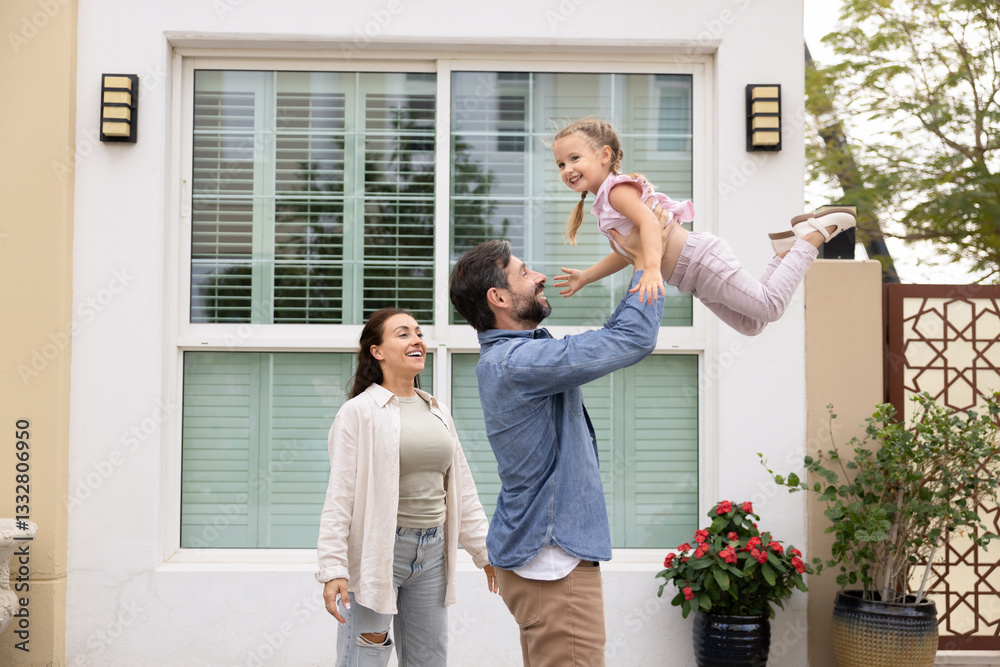 Happy family couple playing with little kid in front of new house. Loving dad lifting cheerful little daughter up in air. Parents and child enjoying homeownership, activity on yard Happy family couple playing with little kid in front of new house. Loving dad lifting cheerful little daughter up in air. Parents and child enjoying homeownership, activity on yard
