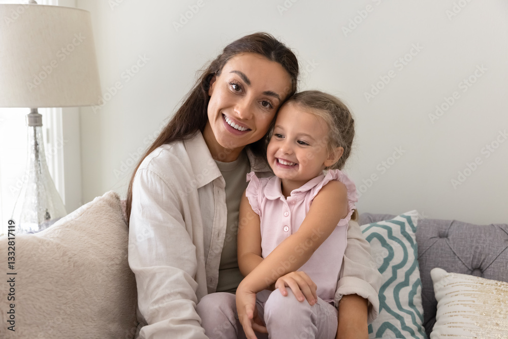 Happy beautiful young mom holding sweet daughter kid on lap, relaxing on comfortable home sofa, enjoying family leisure, motherhood, looking at camera, smiling, laughing, posing for portrait Happy beautiful young mom holding sweet daughter kid on lap, relaxing on comfortable home sofa, enjoying family leisure, motherhood, looking at camera, smiling, laughing, posing for portrait