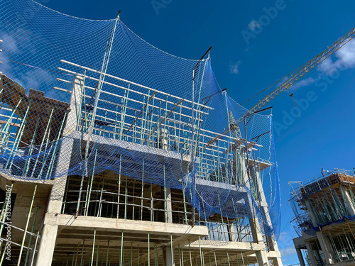 View of the construction site of an apartment building with cranes and safety nets at work, with blue sky and no people working.