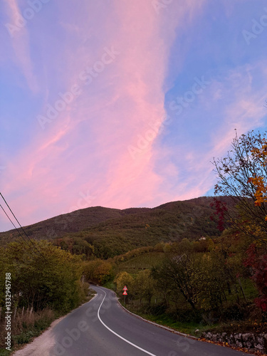 A scenic road in Serbia stretches into the distance between lush green hills, in the warm pink glow of the setting sun on the sky