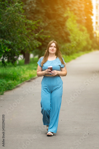 Wallpaper Mural A young woman in a blue outfit strolls through a vibrant park, sipping coffee under golden sunlight. Her relaxed pose reflects a joyful, nature-inspired daily routine. Torontodigital.ca