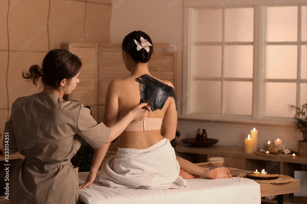 Naklejka premium Young woman undergoing mud treatment in dark spa salon, back view