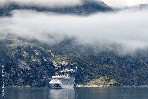 A cruise ship navigates through the stunning Alaskan landscape, surrounded by towering cliffs and foggy skies that create an aura of tranquility and adventure.