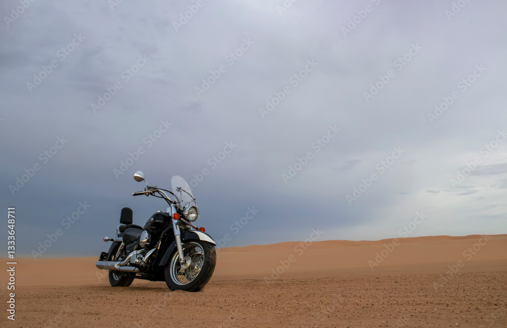 A lone motorcycle stands on the sandy surface of the Namib Desert against a dramatic cloudy sky. The sunset lighting highlights the contrast between nature and machine. A lone motorcycle stands on the sandy surface of the Namib Desert against a dramatic cloudy sky. The sunset lighting highlights the contrast between nature and machine.