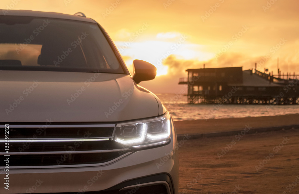 Fototapeta premium A close-up of a gray car parked on the seashore at sunset. The soft light highlights its silhouette, creating a sense of solitude and freedom against the calm waves and vibrant sky.