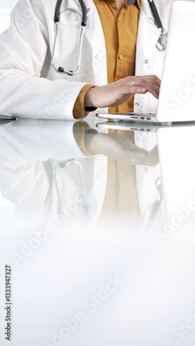 Obraz na płótnie Unknown doctor man in a yellow shirt under a medical coat is using technology in medicine. Close-up of hands typing on a laptop on a glass desk in the clinic, vertical view. Health care concept w salonie