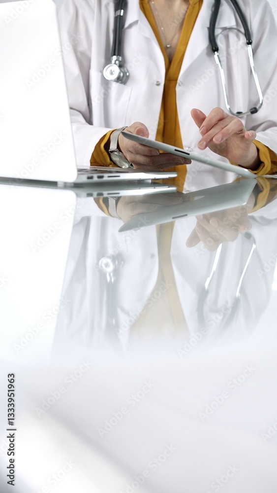 Unknown doctor woman wearing a stethoscope, yellow blouse under a lab coat, is using digital tablet in modern healthcare setting, interacting with patient data, vertical portrait. Medicine concept
