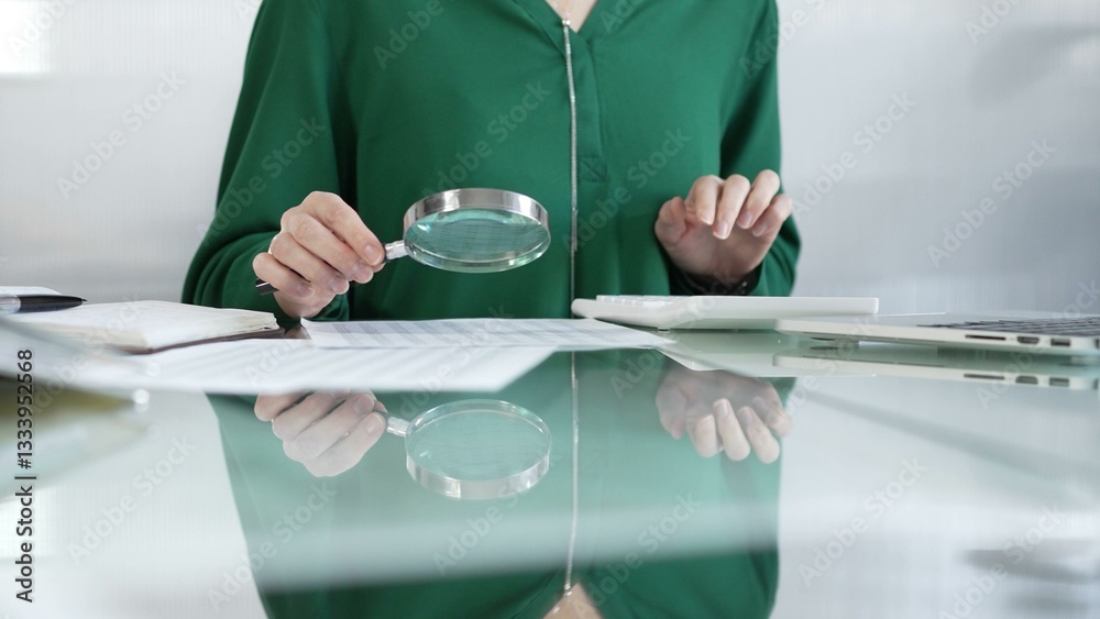 Businesswoman in green blouse carefully reviewing financial reports with magnifying glass and calculator, conducting audit and analyzing data in office. Taxes, audit in business