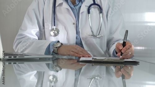 Obraz na płótnie Woman doctor, wearing a blue blouse under a white medical coat, is using a clipboard and taking notes on glass desk in a medical office, close-up. Medicine w salonie