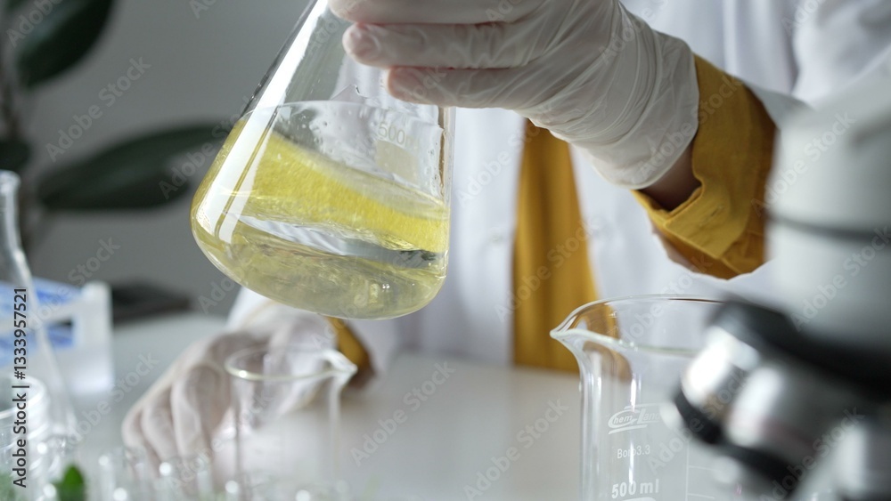 Unknown scientist wearing white protective gloves is carefully shaking up a yellow chemical solution inside an Erlenmeyer flask in a laboratory, close up. Science and medicine