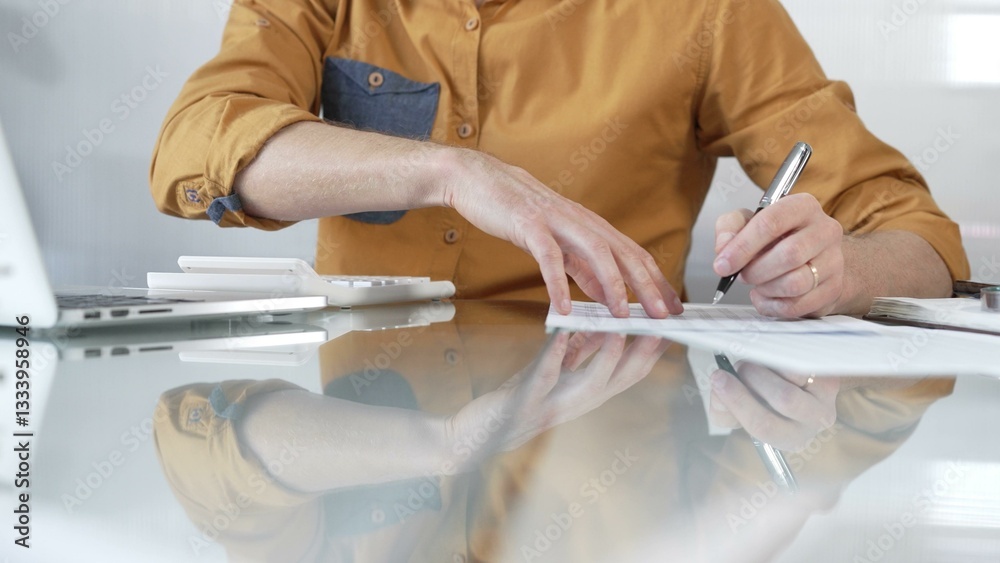 Businessman accountant wearing casual yellow shirt carefully reviewing financial reports using a calculator on office glass desk. Taxes, audit in business