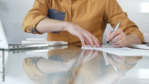 Businessman accountant wearing casual yellow shirt carefully reviewing financial reports using a calculator on office glass desk. Taxes, audit in business
