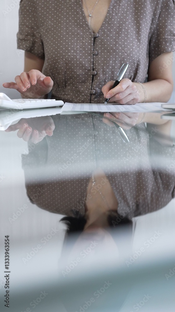 Businesswoman accountant, wearing brawn dotted blouse, is using a calculator at sleek glass desk in a contemporary office, vertical view. Taxes, audit in business