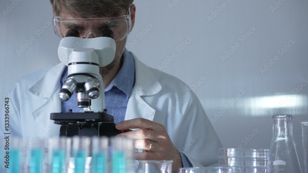 A man scientist, wearing a lab coat, mask, and safety glasses, is using a microscope surrounded by test tubes in laboratory setting. Medicine and science concept
