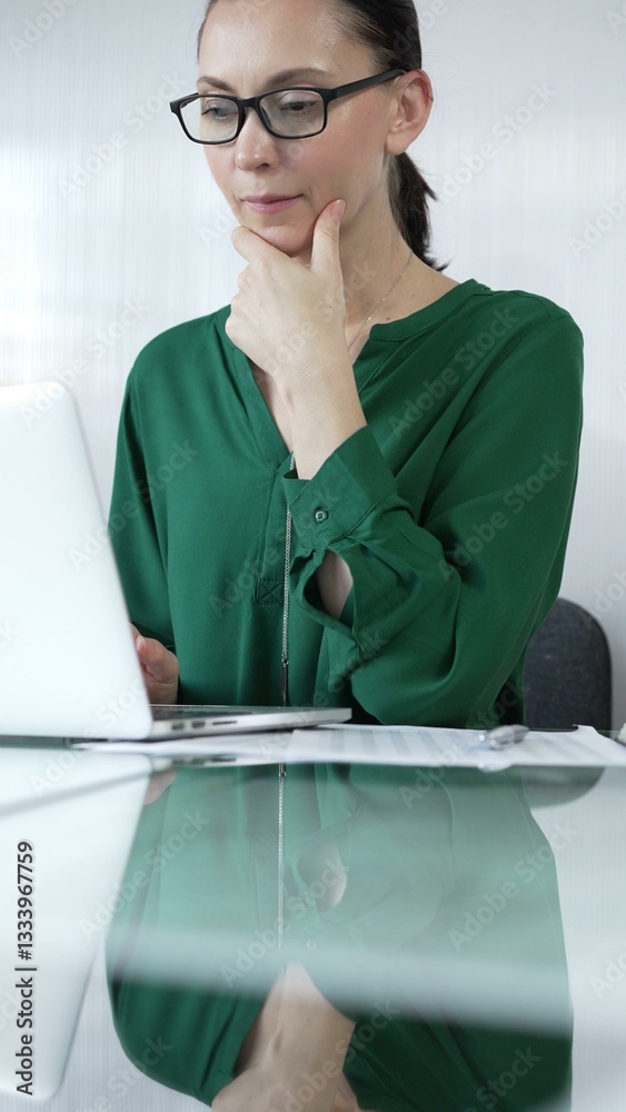 Focused businesswoman in green blouse is using laptop and thinking with hand on chin, working on a project in a bright office, vertical portrait. Business people concept