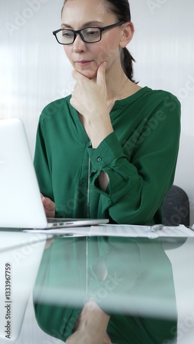 Obraz na płótnie Focused businesswoman in green blouse is using laptop and thinking with hand on chin, working on a project in a bright office, vertical portrait. Business people concept w salonie