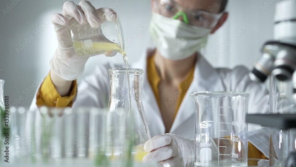 Female scientist researcher wearing a lab coat, white gloves, and mask, is pouring a yellow oily liquid from a beaker into an Erlenmeyer flask in laboratory, portrait. Science and medicine concepts