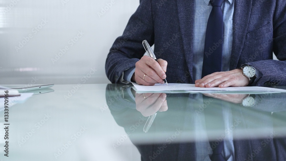 Businessman in a professional attire signing a contract with a pen on a modern glass desk in a contemporary office environment. Business people or audit concept