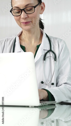 Obraz na płótnie A female doctor in a green blouse under a medical coat is using technology in healthcare. Woman is typing on a laptop on a glass desk in the clinic, vertical portrait view. Medicine w salonie