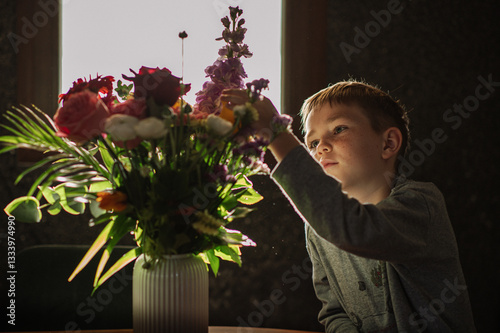 A little boy admires a bouquet of flowers in a vase on the table at home, gently touching the petals with his finger and enjoying the fragrance.