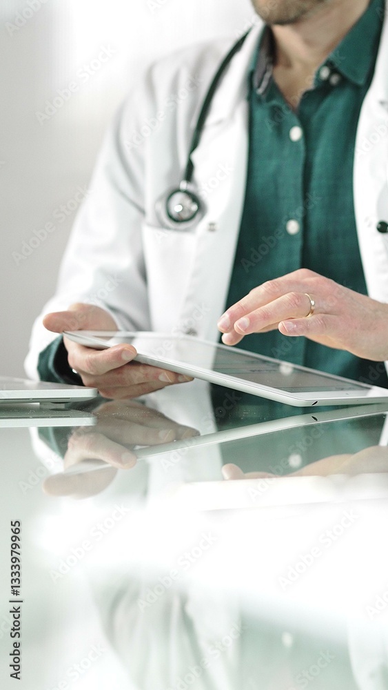 Doctor man in green shirt under a white coat using digital tablet in modern healthcare setting, interacting with patient data. Medicine concept