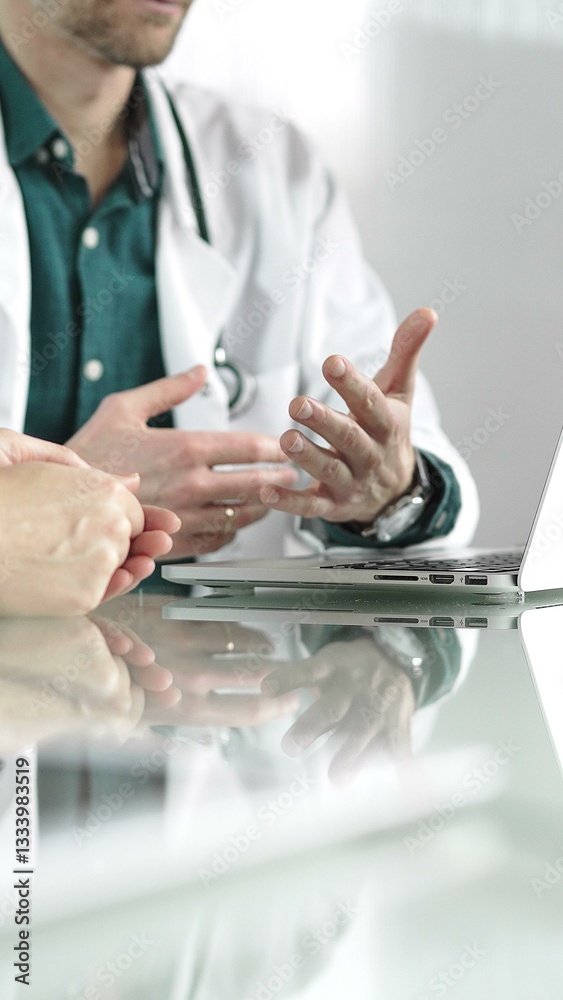 Doctor man wearing a stethoscope and lab coat is gesturing with hands while explaining a diagnosis to a patient across a desk with a laptop. Medicine concept