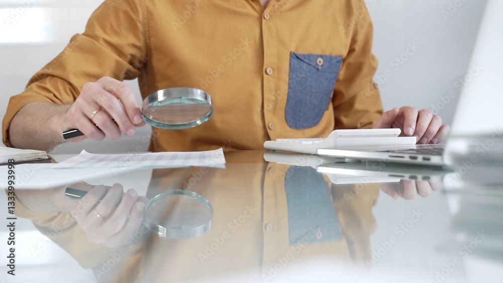 Businessman accountant wearing casual yellow shirt carefully reviewing financial reports with magnifying glass and calculator, conducting audit and analyzing data in office. Taxes, audit in business