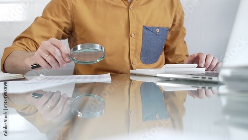 Obraz na płótnie Businessman accountant wearing casual yellow shirt carefully reviewing financial reports with magnifying glass and calculator, conducting audit and analyzing data in office. Taxes, audit in business w salonie