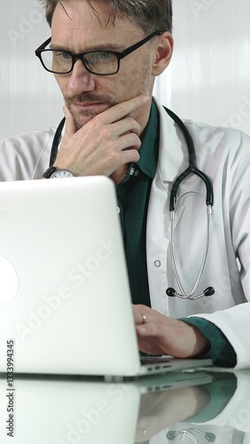 A male doctor in a green shirt under a medical coat is thinking while using technology in healthcare. Man is typing on a laptop on a glass desk in the clinic, vertical portrait view. Medicine concept