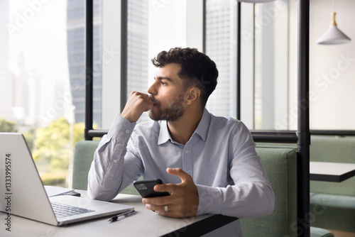 Pensive, thoughtful young male freelancer sit at desk with laptop in modern coworking office holding phone waiting for response, reviewing received information, or considering work or personal matter